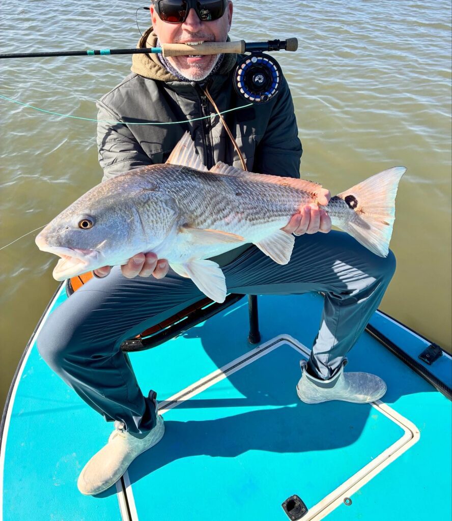 Man in outdoor gear holding a freshly caught fish by the water.