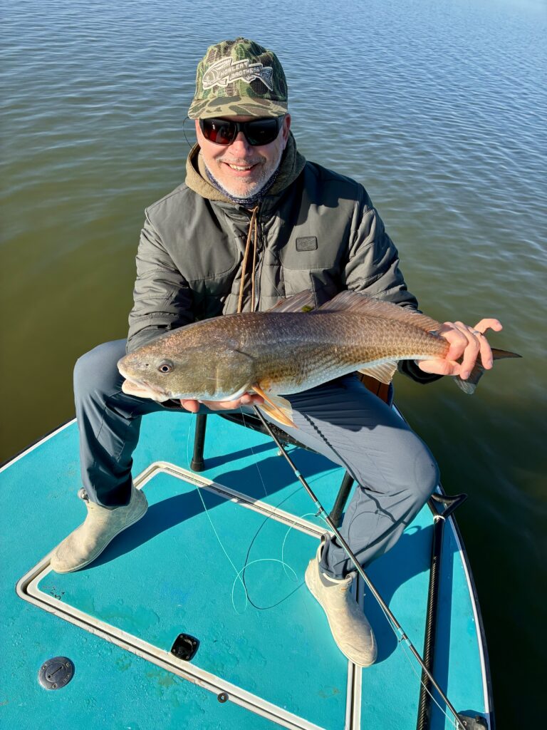 Man in outdoor gear holding a freshly caught fish by the water.