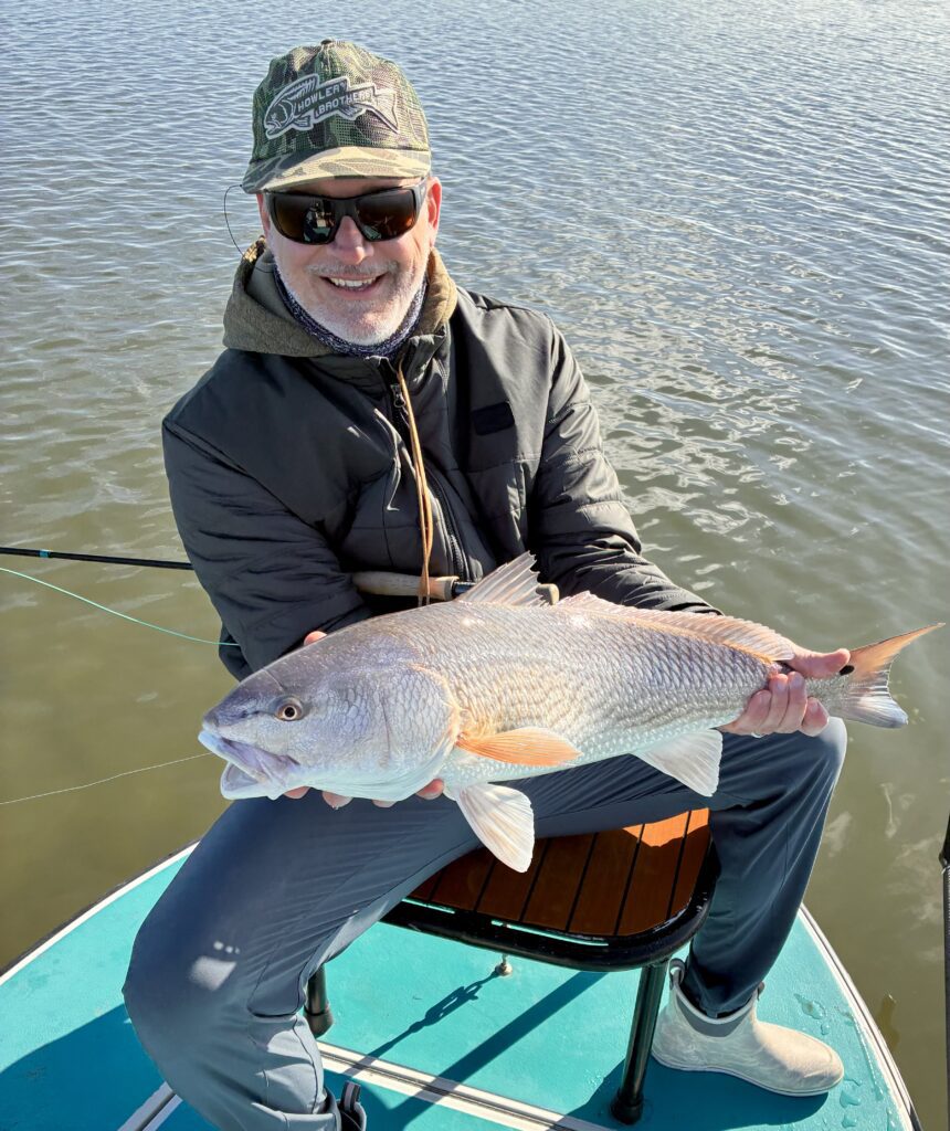 Man in outdoor gear holding a freshly caught fish by the water.
